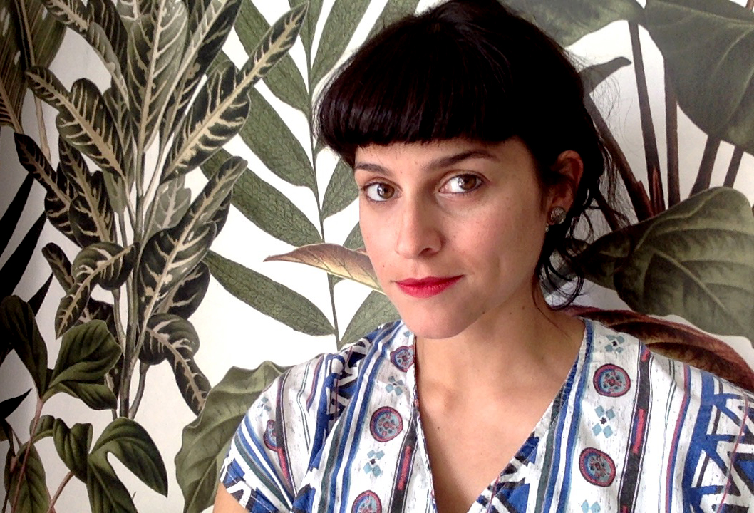 Woman with dark hair has a slight smile, wearing a white /blue patterned shirt. Behind her is a green leafy patterned wallpaper.