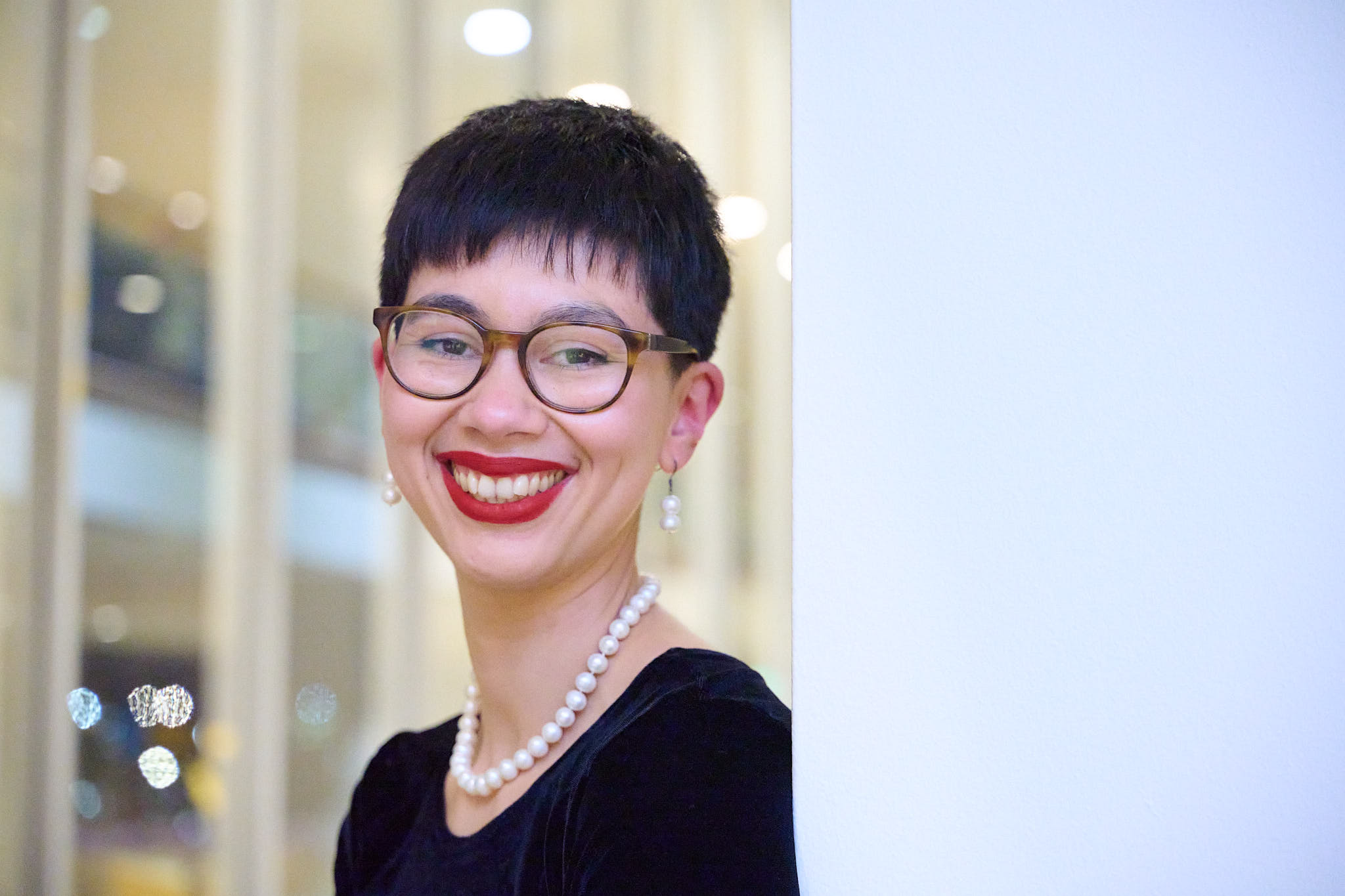 Helen Bowell smiles towards us. She has short black hair and wears a black velvet top, pearls, red lipstick and dark glasses.