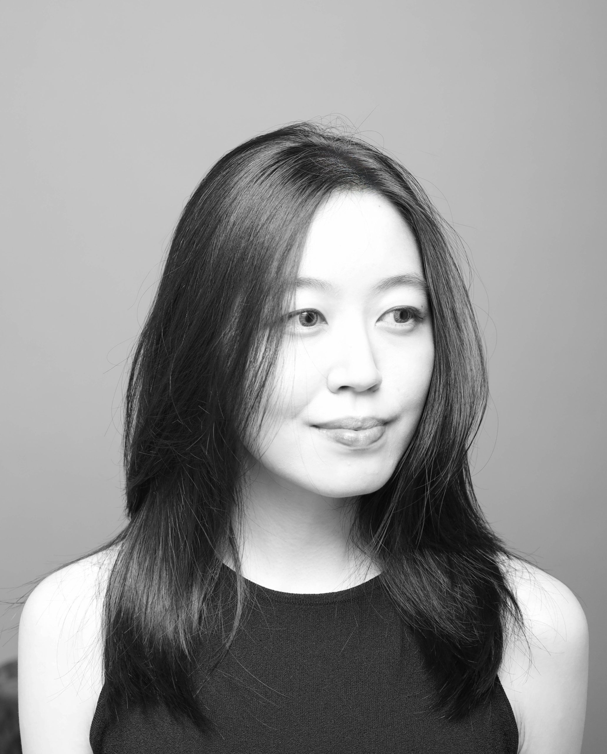 A black and white photo showing an Asian woman with long black hair. Her head is slightly turned to her left and she is looking off-camera. She is wearing a black top and behind her is a plain background.