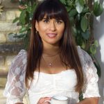 An Indian woman stands in front of a yellow brick wall, half of which is covered in green, crawling hoya leaves. She has long dark hair, which is worn straight, a fringe, and brown eyes. She smiles at the camera in a white lace blouse and blue jeans, whilst holding a light blue mug of tea.