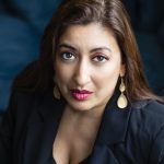 Centre-frame, an olive-skinned woman stares up into the camera. The background is dark and moody. She has long, brown hair and wears a blue blazer.