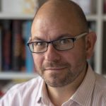 Head shot of a bald, middle aged light skinned man in glasses against a backdrop of a bookshelf.