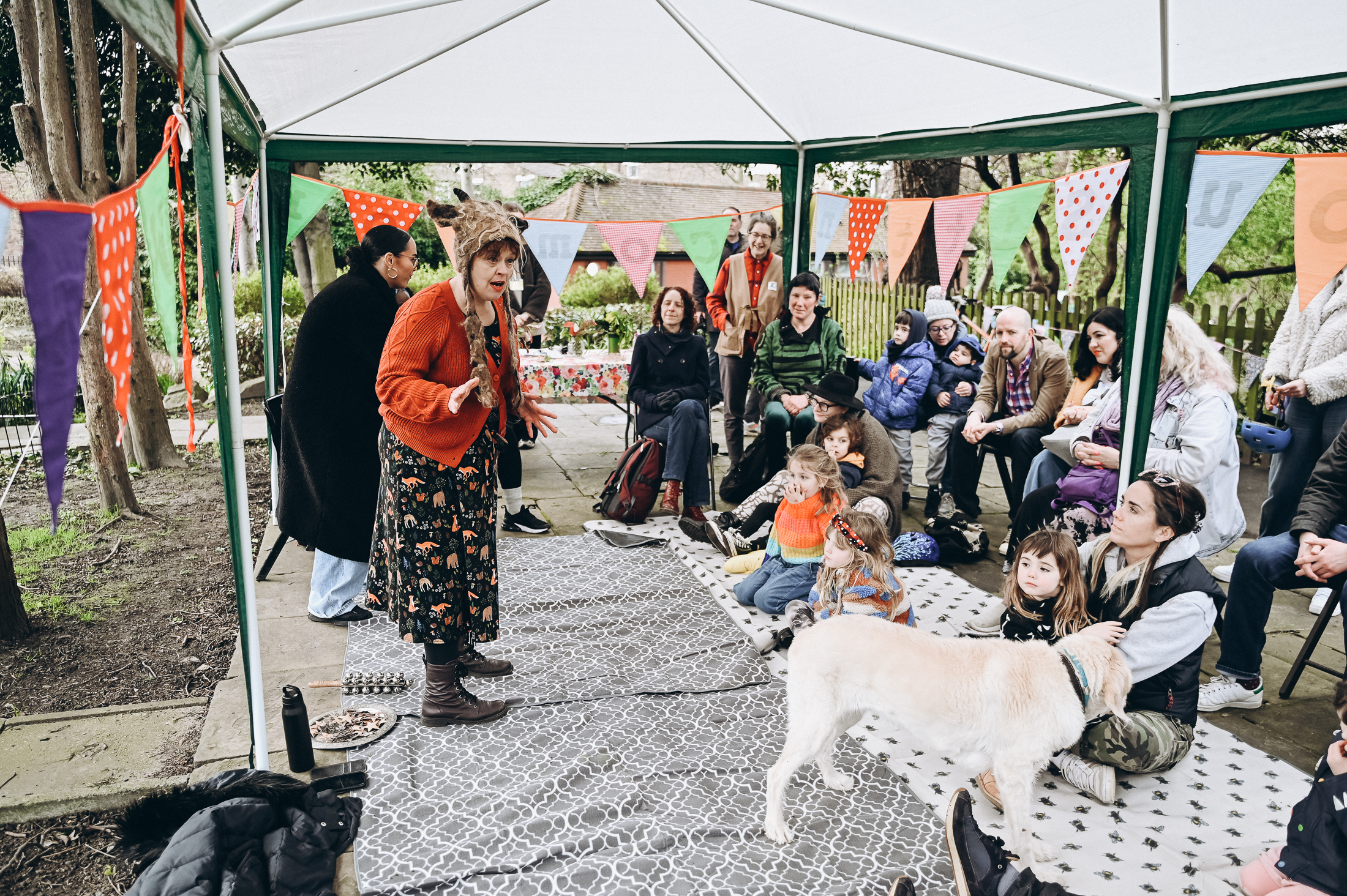 Under a gazebo in a park, families sit on cushions while two performers tell them a story.