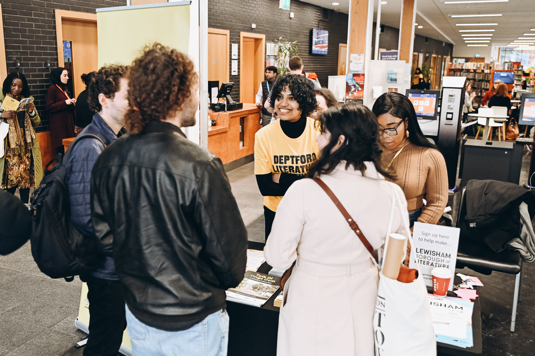 In a library, a person with a bright yellow t-shirt reading Deptford Literature Festival stands behind a table, speaking to some people on the other side of the table.